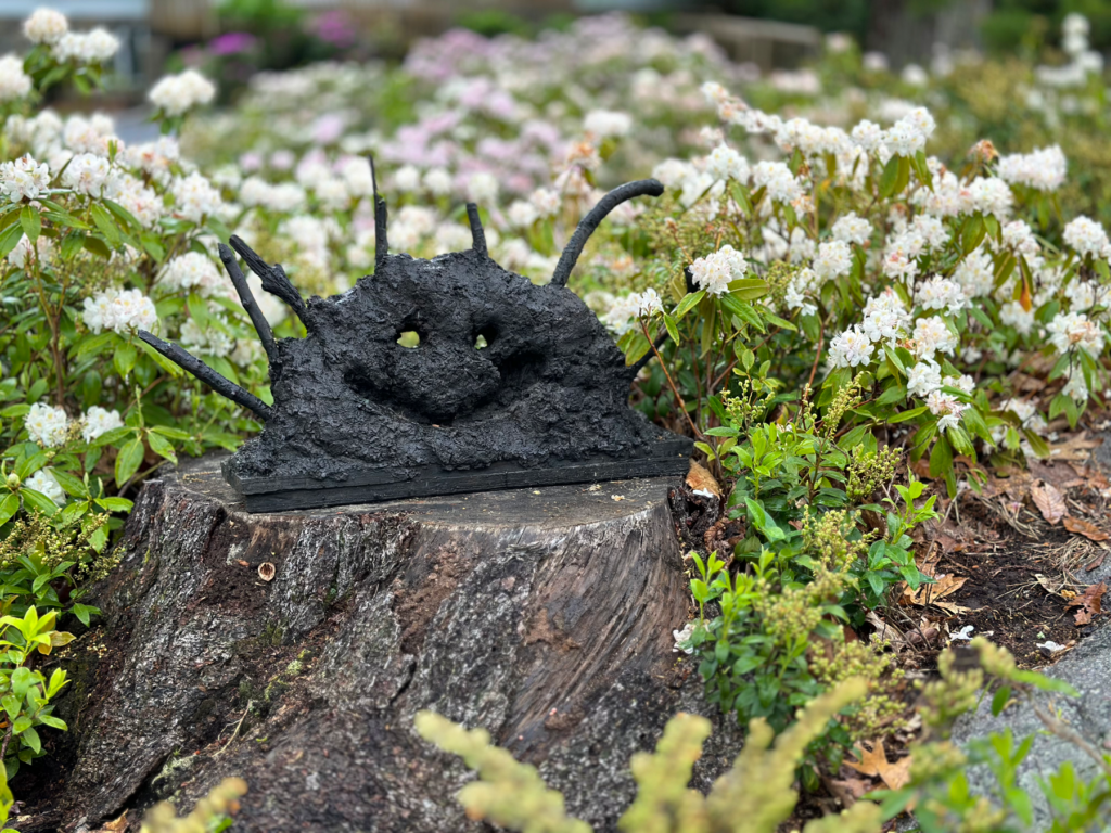 Photo of a bronze sculpture by artist Patrick Jacobs, entitled "Smiling Head" installed in a bed of Azaleas that are in bloom